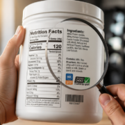 A high-resolution, photorealistic close-up shot of a person's hands holding a white plastic protein supplement container with a detailed nutrition label facing the camera. The label is in sharp focus, showing protein content, ingredient list, serving size, and third-party certification logos. A magnifying glass hovers over the label, emphasizing the act of careful reading. Soft natural lighting from the left, shallow depth of field with a blurred gym or kitchen background in warm neutral tones. Clean, modern editorial photography style, vertical composition suitable for a blog header, ultra-detailed, 4K quality.