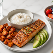 A high-quality, photorealistic flat-lay composition on a clean white marble countertop featuring a balanced refeed meal: a grilled salmon fillet, a bowl of jasmine rice, roasted sweet potatoes, fresh avocado slices, mixed berries, and a glass of water. Soft natural daylight from the left, shallow depth of field, minimalist aesthetic, subtle metabolic and hormonal health theme, no text, editorial food photography style, 4K, ultra-sharp details, warm tones.