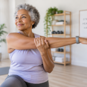 A serene, well-lit image of an active senior woman (around 65-70 years old, diverse ethnicity) performing a gentle stretching exercise (e.g., arm raises or leg lift) with a calm, focused expression. She is wearing comfortable athletic wear in a bright, modern home gym or a clean, minimalist studio. The background is slightly blurred, suggesting a peaceful environment. The overall mood is positive, empowering, and emphasizes safety and well-being. High quality, realistic, natural light.